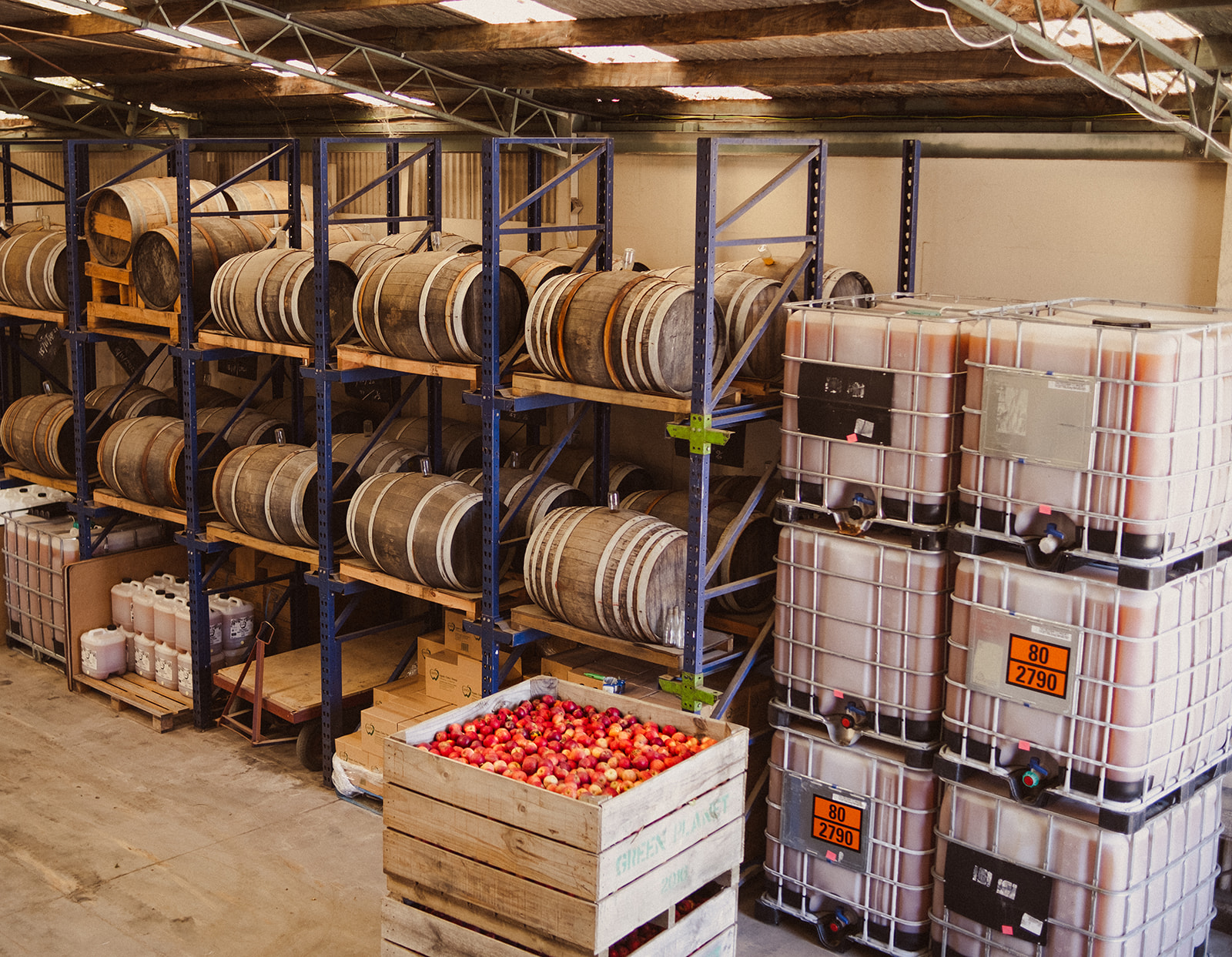Crates of fresh apples sit in the CoralTree warehouse. Behind are oak barrels with aging Apple Cider Vinegar.