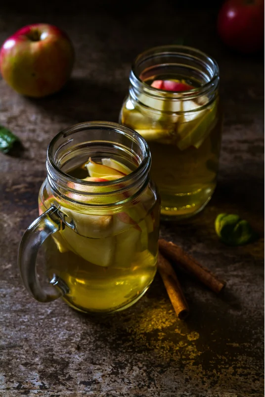 Two glass jars filled with apple cider vinegar, featuring slices of fresh apples and cinnamon sticks, set on a rustic wooden surface.