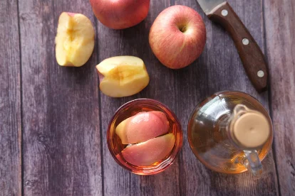 Freshly sliced apples and whole apples next to a glass and jar for apple cider vinegar preparation.